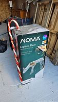 Photo showing NOMA Lighted Deer box with candy cane decoration leaning against it on porch floor.