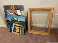 Unframed paintings stacked against wall with two unpainted wooden frames leaning nearby