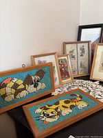 Collection of framed pictures including two vintage mosaic-style cat images and smaller framed art prints arranged on a lace-covered tabletop.