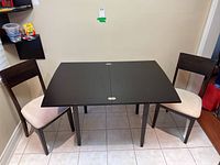 Front view of black wood kitchen table with flip-top and two beige upholstered chairs positioned on either side against a beige wall.