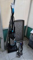Photo of the Bissell Powerforce upright vacuum cleaner with black and blue handle, transparent dust container, and power cord wrapped around the back. Behind it is a decorative gray boot mat with an embossed scroll pattern.