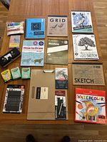 Overview of all items laid out on wooden floor including sketch pads, pens, crayons, markers, pencils, and art books, all unused in original packaging.