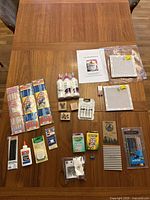 Lot overview on wooden table showing all items including pipe cleaners, glue, stamps, needlepoint canvases, pencils, notepads, crayons, Rick rack trims, tape measure and sewing pins.
