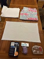 Overview of all items on wooden table: 2 white crochet-trim bath rugs, blue and pink patterned shower curtain folded, box of Philips Sonicare toothbrush, purple travel soap container, clear toiletry bag.
