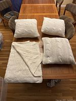 Four items displayed on wooden table: one folded beige cable-knit throw blanket and three matching knit decorative square pillows.