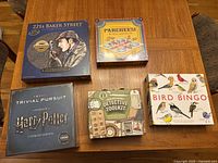 Overview of all five sealed board games on a wooden table showing box art for Sherlock Holmes 221B Baker Street, Parcheesi Royal Edition, Harry Potter Trivial Pursuit Ultimate Edition, Bird Bingo, and Sherlock Holmes Detective Toolkit.