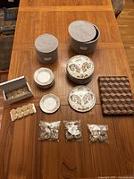 All items shown on wooden table: 8 small and 8 medium plates in grey fabric containers, wooden cheese board with checkered pattern, boxes of beeswax candles, gold napkin rings in bags
