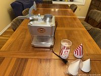 Front view of Waring snow cone maker with lever and clear plastic bin. Paper and plastic cones placed beside it.