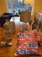 Photo showing two glass base lamps with rope net design and burlap shades on a wooden table with two seaside-themed pillows folded beside them