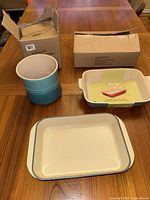 Three Le Creuset cookware items displayed on a wooden table with their original boxes: the utensil crock, ceramic dish, and cast iron dish.