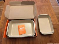 Two rectangular cast iron baking dishes, one larger in box, one smaller used beside it, showing interior underwing beige enamel and green exterior.