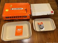Two red Le Creuset rectangular roasters (large left, small right) with original boxes behind on table; cream enamel interiors visible; boxes torn