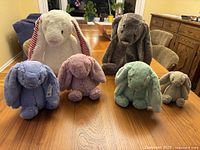 All six Jellycat bunnies arranged on a wooden table—white with red-striped ears, gray, blue, lavender, mint green, blush pink