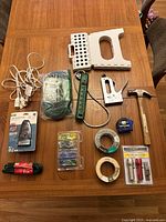 Tools including hammer, staple gun, step stool, power strip, tapes, measuring tape, and bit set laid out on a wooden table.