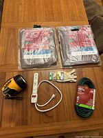 Two new packaged brown 8x10 tarps, a new green 50 ft outdoor extension cord, new women's garden gloves, a white 6-outlet power strip, and a yellow and black Rayovac industrial krypton flashlight/lantern on wood table.