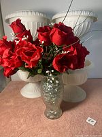 Red artificial flower arrangement in a cracked-texture glass vase with white baby's breath and decorative grass, placed on a pink surface with white urns in the background.