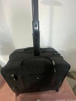 Side front view of the black rolling briefcase on a pink surface showing telescoping handle extended and zippered compartments.