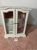 Front view of the white wooden jewelry organizer cabinet with floral etched glass doors, showing interior compartments and hooks.