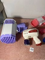 Photo showing three toy bubble guns and two bottles of bubble solution on a carpeted floor against a white wall.
