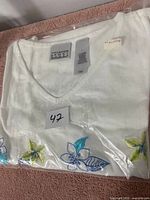 White ladies T-shirt with floral embroidery visible, folded in plastic packaging with size and brand tags showing.
