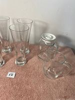 Six tall clear glass drinking glasses arranged on a towel surface with a clear glass jug and a clear glass jar with a white plastic lid.