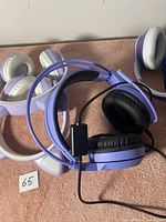Photo showing multiple pairs of over-ear headphones in light purple with black ear pads, white with purple accents, wired with inline control.