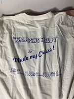 Back view of white T-shirt with large blue text 'SHOPPING SHELBY Made my Cruise!' and 'I SAW IT ...... I LOVED IT ...... I BOUGHT IT ......'.