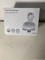 Photo showing front of the product box, featuring the image of the white scalp massager and icons indicating touch job, waterproof, and exchange patterns.