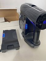 Cordless vacuum main body and extra battery both showing LED battery charge indicators, placed on a white surface with a cardboard box behind.