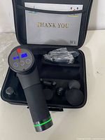 Wattne1 cordless massager with digital display, controls, and visible massage head, placed inside open case with mesh pocket holding instruction booklet.