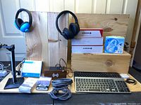 Photo showing blue and black wireless headphones, two boxed Argon HDMI cables, a blue and white earbud set, wireless keyboard, and other accessories on a wooden display.