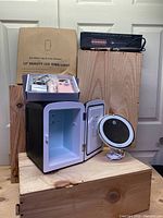 Overview of beauty and hairstyling bundle showing the makeup mini fridge with door open, LED makeup mirror next to it, packaged 12-inch LED ring light box behind, and electric nail drill in its box on top of the fridge.