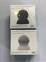 Two unopened boxes of Mission Battery Bases for Amazon Echo Dot 4th Gen stacked vertically. The boxes show different color depictions of the Echo Dot on each base.