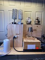 Front view of seven decorative lamps arranged on wooden boxes including cylindrical chrome base lamps with lattice metal shades, geometric lantern-style lamps, and a silver bell-shaped desk lamp.