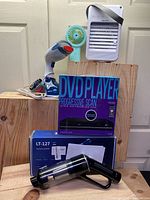 Five items arranged on wooden blocks: a handheld garment steamer, a blue handheld fan, a white and black air cooling fan, a boxed Proscan DVD player, and a black handheld vacuum cleaner.