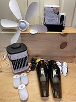 Cooling fan with five white blades, box with essential oil diffuser, white electronic pest repellers, two black handheld vacuums, and white heating or cooling device.