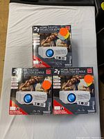 Three sealed boxes of RCA Home Theater Projector Bundles arranged on a fabric surface, showing front packaging.