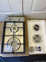 Top view of new stainless steel Ramblewood 2 burner gas cooktop with protective plastic covering on burners and knobs, showing black cast iron grate and two control knobs.