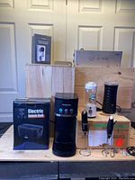 Photo showing the full set of kitchen appliances arranged on a table with packaging including the electric lunch box box visible, and individual items like coffee maker, milk frother, vacuum sealer, paper towel holder, and hand blender attachments.