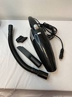 Black handheld vehicle vacuum with power cord coiled beside it, placed on a white surface. Accompanied by three attachments: flexible hose, crevice tool, and brush nozzle.