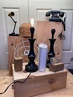 Photo showing the two vintage black metal nightstand lamps with candlestick bases, a wicker lighting fixture, and the antique style black rotary phone on layered wooden blocks.