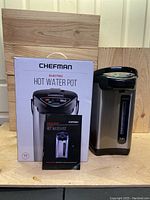 Photo showing the Chefman electric hot water pot standing next to its retail box and user manual. The pot is stainless steel with a black top and base, featuring water level gauge and control buttons.