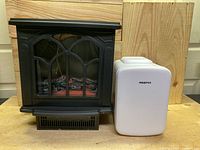 Photo showing black electrical fireplace heater and white thermoelectric cooler side by side on wooden surface with wooden background.