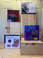 Photo showing all items in original boxes arranged on wooden boxes against wall. Boxes include two radio cassette boomboxes and two portable DVD players (7 and 9 inch).