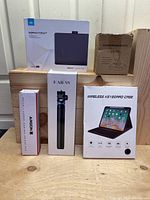 Lot items arranged on wooden surface including Huion Inspiroy Frego2 tablet box, KAESS tripod box, wireless keyboard case box, portable mouse pad, and phone holder boxes.