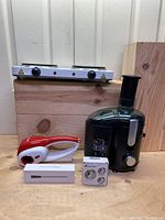 Full lot showing black electric juicer, white and red electric cheese grater with two drums, double burner white hot plate, wireless thermometer box, and kitchen timer.