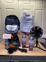 Lot contents showing black backpack, white folded air mattress, two insulated tumblers, rechargeable fan with tripod stand, and headphones with other small items.