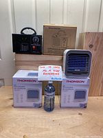 Photo showing professional ozone generator next to its cardboard box, two boxed Thomson mini portable air conditioners, and one unboxed air conditioner on top of the boxes with a water bottle for scale.