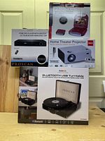 Photo of all items boxed and stacked: Proscan Bluetooth USB Turntable, Proscan HDMI DVD Player with USB port, RCA Home Theater Projector, and 7-inch portable DVD player with red lid.