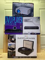 Photo showing the four boxed items: RCA home theater projector, Bluetooth USB turntable, and two Proscan DVD players with progressive scan